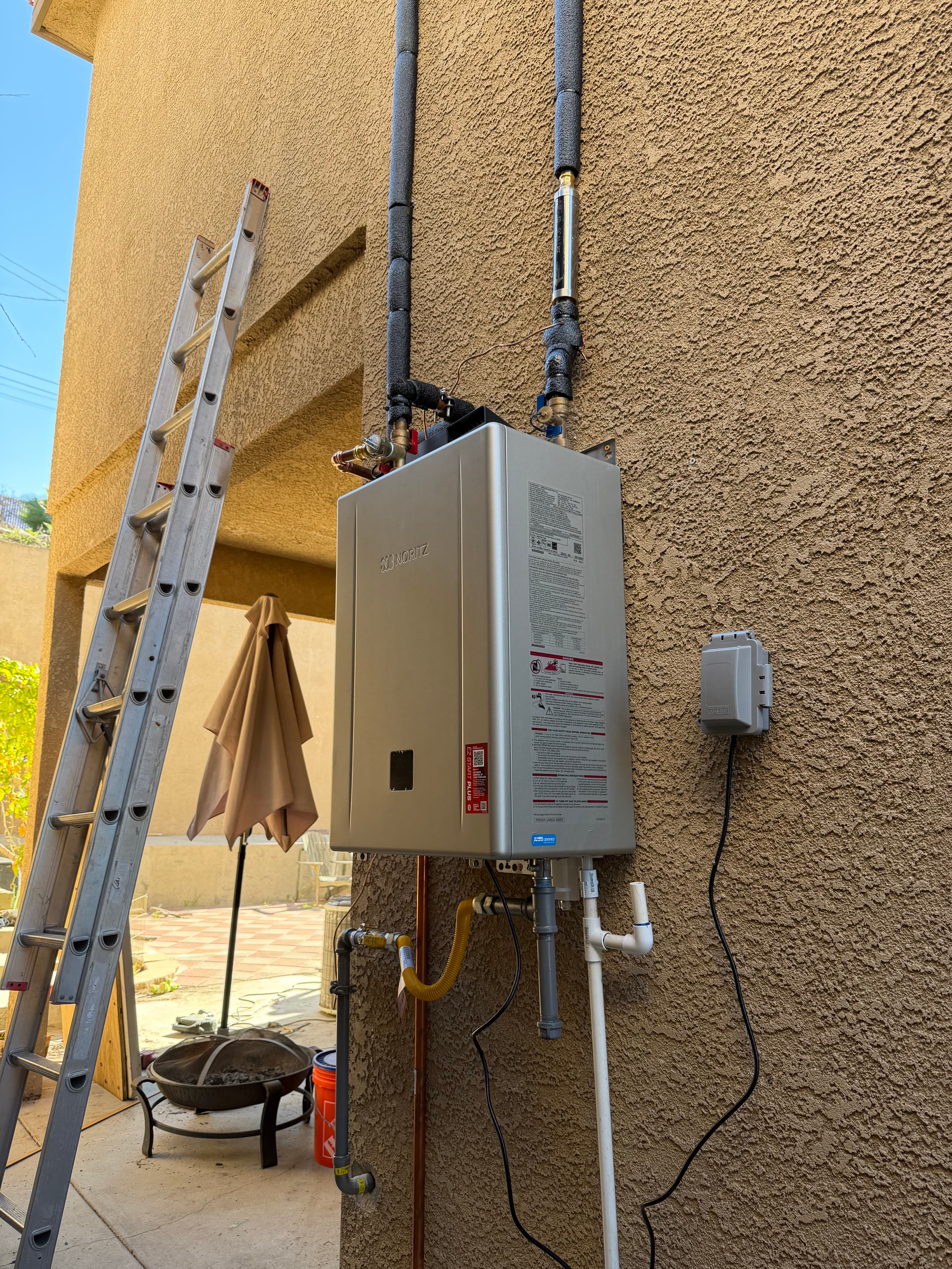 Silver Noritz tankless water heater installed on an exterior stucco wall next to a ladder.