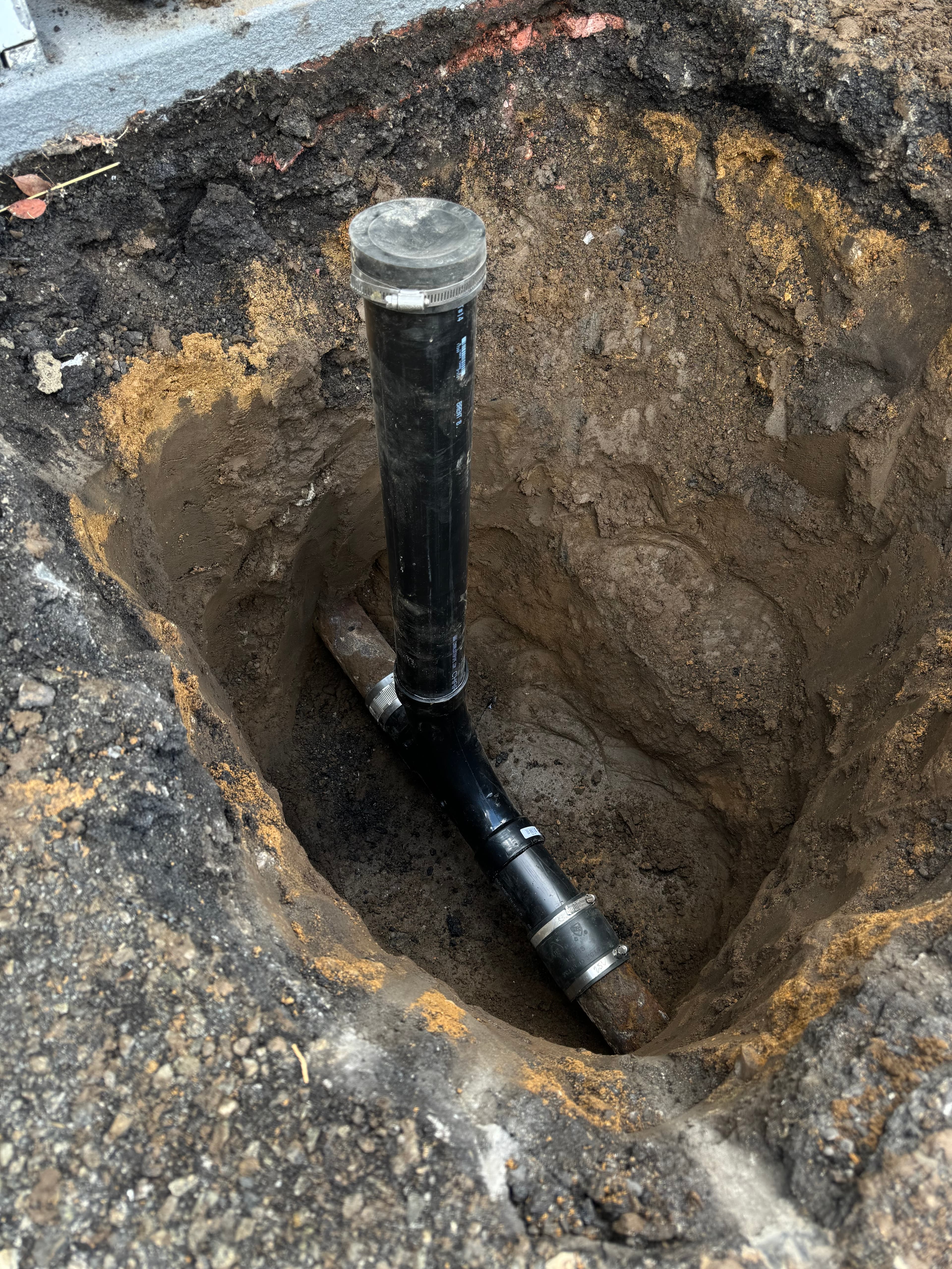 Vertical black PVC cleanout pipe connected to underground sewer lines within a deep dirt hole.