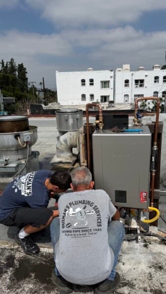 Two plumbers from Guzman's Plumbing Services install a tankless water heater on a rooftop.