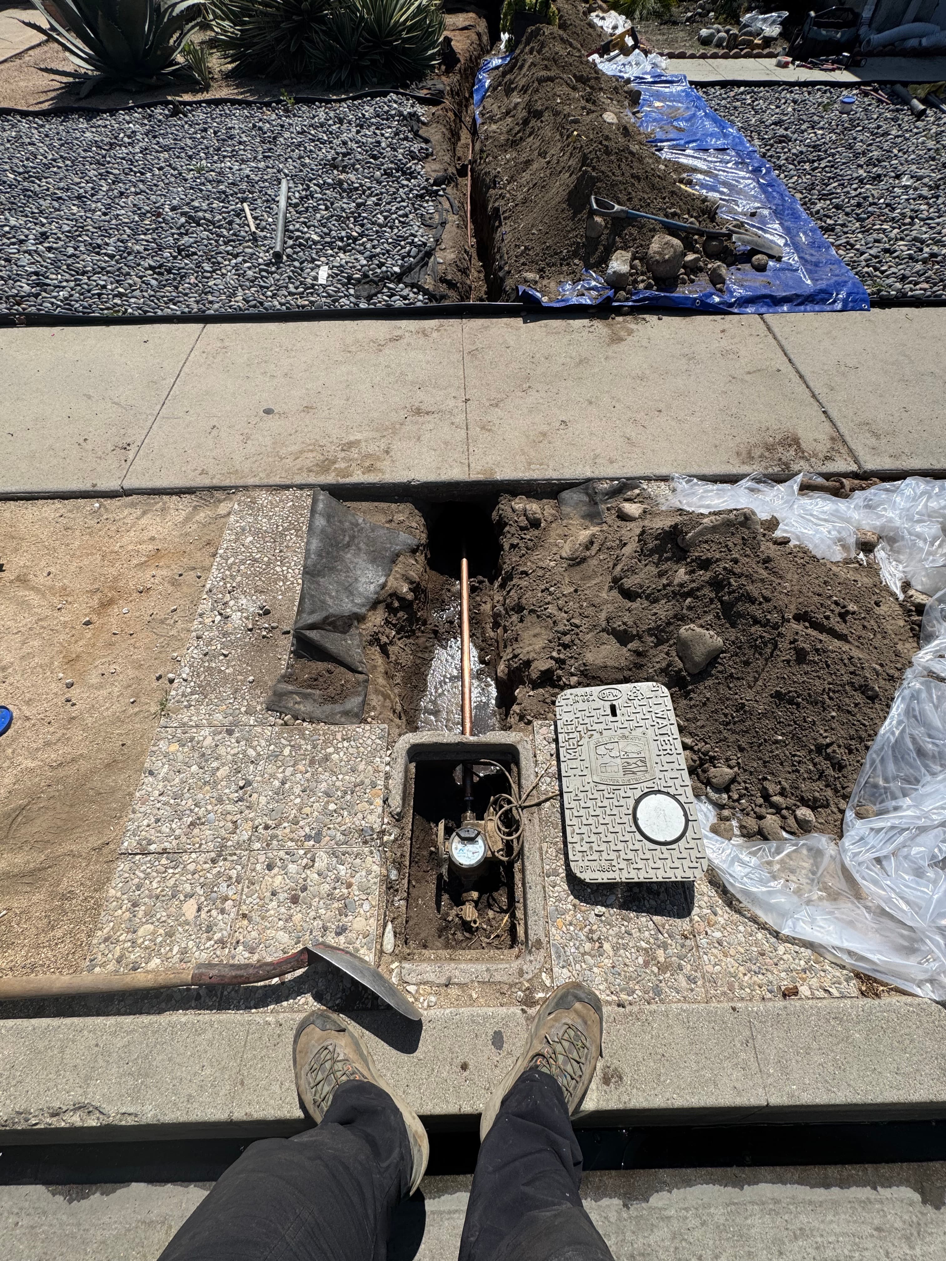 Trench dug across a sidewalk for water line repair with an open meter box.